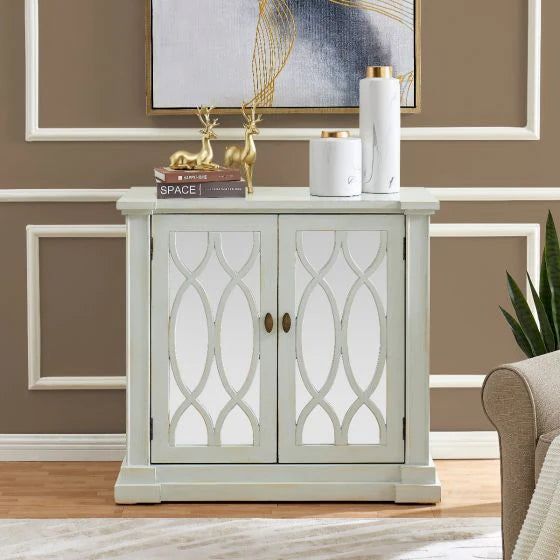 A Modena Small Sideboard with mirrored glass panels and gold handles is topped with gold deer figurines, books, and white vases. The sideboard is set against a taupe-brown wall accented by crisp white trim.