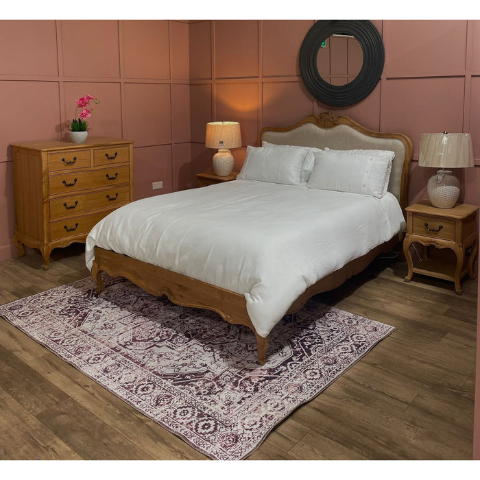 A cozy bedroom with a French style Toulouse oak bedframe and upholstered headboard, white bedding, solid oak nightstands with lamps, a matching dresser, round wall mirror, orchid plant, and patterned rug on wooden flooring.