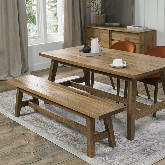 A modern dining room features an oak table, Camden Bench, and two orange mid-century armchairs. A white rug lies on the wood floor, with large curtained windows and a sideboard completing the inviting setting.