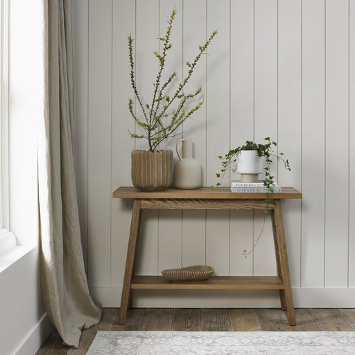 The Camden Console Table with oak tops stands against a light paneled wall, holding three vases with branches and greenery. A ribbed bowl is on the lower shelf, with beige curtains and a patterned rug partially visible.