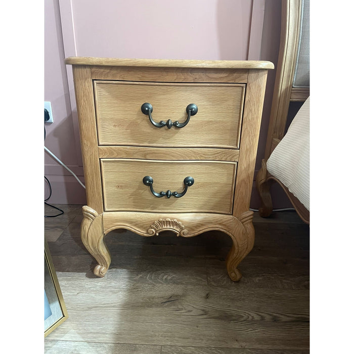 The Toulouse Bedside Locker, with two drawers, ornate handles, and curved legs, stands on a wooden floor beside a pink wall and a partially visible bed, adding charm to this French style oak bedroom.