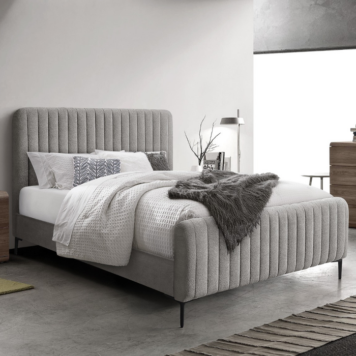 A modern bedroom with the Clarissa Bedframe in grey fabric and a vertically lined headboard, white and gray bedding, a small wooden nightstand, black floor lamp, decorative branches in a vase, all in a minimalist contemporary style.