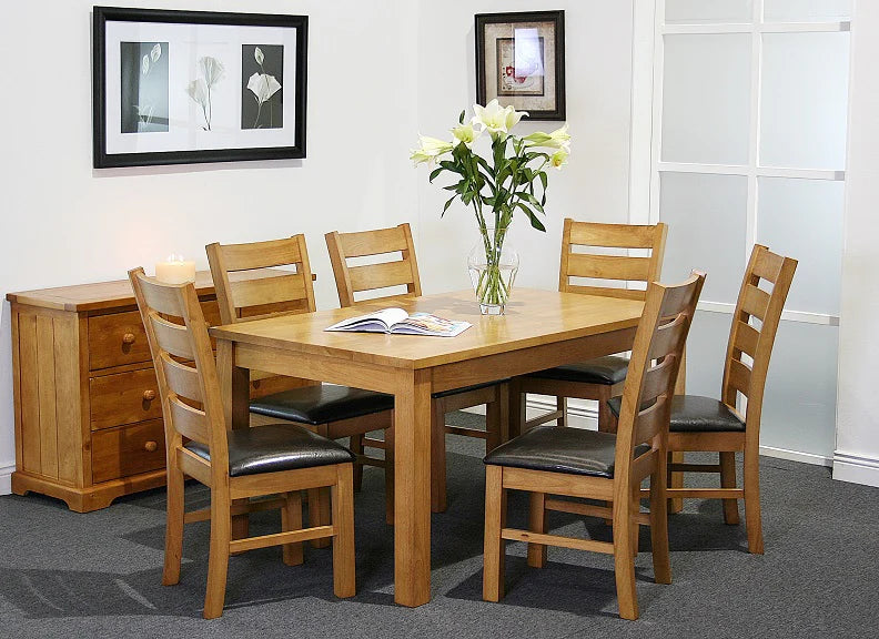 The Columbia Dining Set - Oak, with six padded chairs, surrounds a vase of white lilies and an open book. A candle-topped sideboard stands nearby, while framed art adorns the walls of this carpeted room.