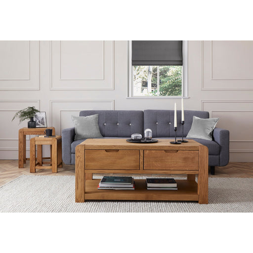 Modern oak coffee table with soft rounded corners and visible wood grain, placed in a room with a grey sofa, on a white carpet.