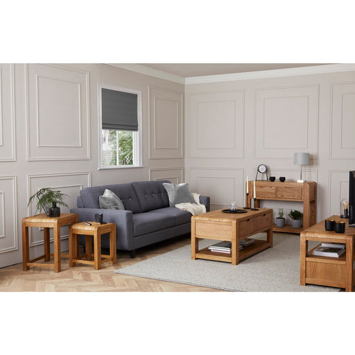 A modern living room features a gray sofa, light paneled walls, a window with a gray blind, and the Edson Nest Of Tables in solid oak on a beige rug, complemented by minimalist decor, cushions, lamps, and an oak sideboard and coffee table.