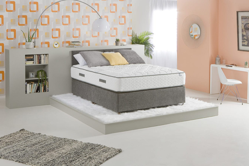 A King Koil Paradise mattress displayed in a modern bedroom setting, showing the top surface with pillows on it, encased in a gray and white cover.