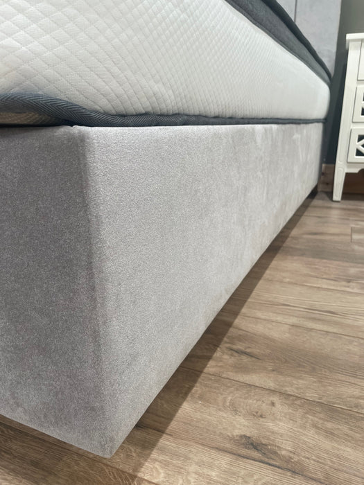 Close-up of the Bella Ottoman Bed in light gray upholstery with a white mattress on top, set on a wooden floor beside a white nightstand.
