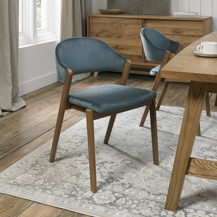 A modern dining room featuring a wooden table, two Camden Dining Chairs in blue velvet with wooden legs, a patterned rug, wooden sideboard, and a large window adorned with beige curtains.