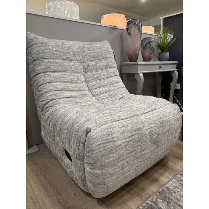The Ryder Occasional Chair, featuring plush grey fabric and a curved design, sits on a wooden floor beside a small gray table topped with two decorative vases and a green plant.