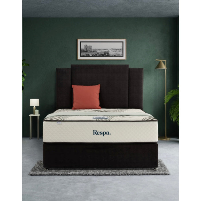 A modern bedroom with a dark upholstered bed, a cream Respa Jupiter mattress, one orange pillow, two side tables with lamps, a floor lamp, a framed picture, and a plant against a dark green wall.