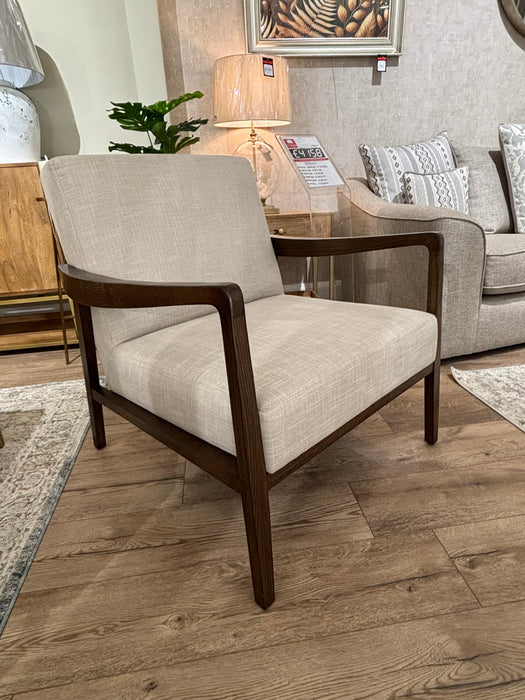 The Monza Armchair, featuring a walnut wood frame and light beige upholstered seat and backrest, is showcased on a wooden floor in a stylish living room display.