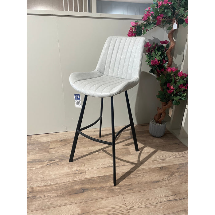 The George bar stool features padded, ribbed gray leather seats and black metal legs. It stands on a wooden floor beside a potted artificial plant with pink flowers.