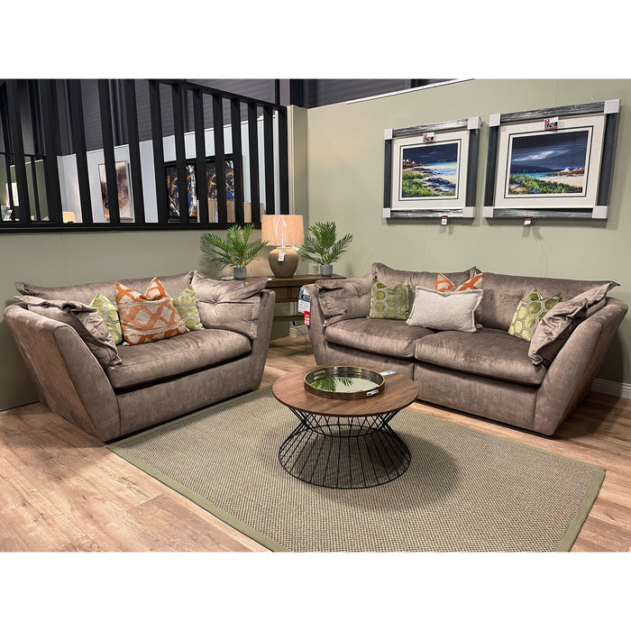 A modern home interior with two Sully taupe sofas, colorful pillows, a round wooden coffee table with wire base, beige rug, table lamp, green plants, and two landscape paintings on a light green wall.