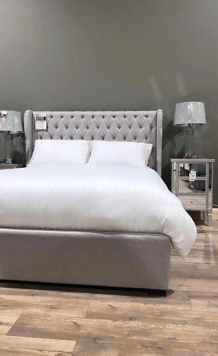 A Vienna Bedframe with a tufted gray winged headboard, dressed in white bedding and two pillows, sits on a wood floor against a muted green wall, flanked by mirrored nightstands with plastic-covered lamps.