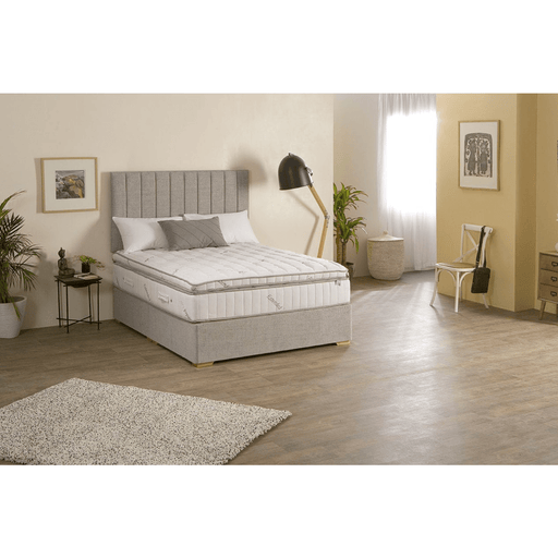 A King Koil Extended Life Plus mattress with a low profile divan, displayed in a bright bedroom setting, featuring a gray upholstered headboard and two-sided reversible box-topper.