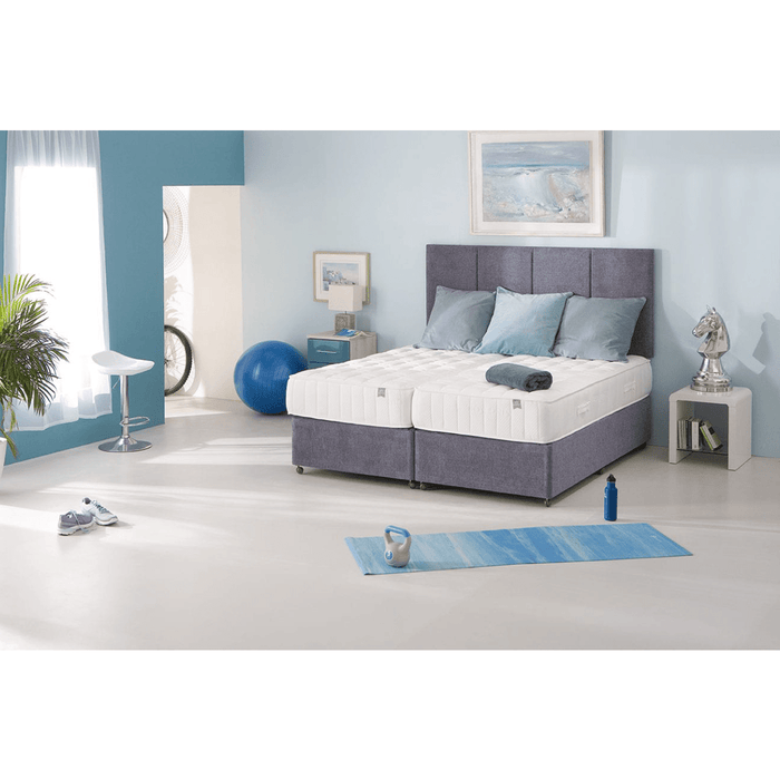 A modern bedroom features a large grey King Koil Spinal Guard (ICA) mattress by vendor-unknown, light blue and white bedding, folded towels, blue walls, fitness gear, a yoga mat, and framed art for a calm space with optimal posture support.
