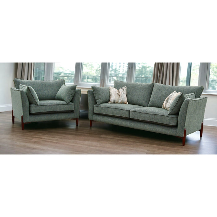 A modern living room features Layla furniture—a gray-green, made-to-measure couch and matching armchair with wooden legs and decorative pillows—set on a wooden floor near windows draped with beige curtains.