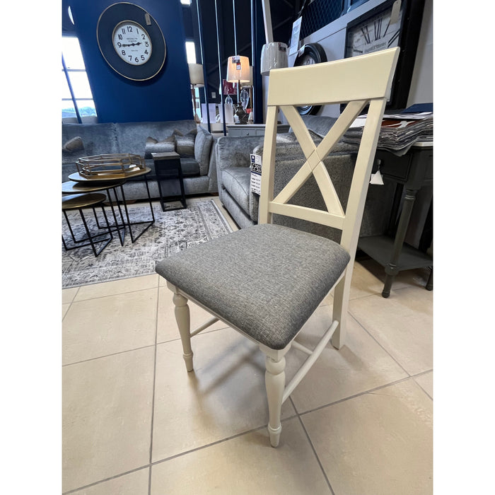 The Angelia dining Chair, featuring a white wooden X-back and a gray cushioned seat, is showcased next to a cream dining table in a modern showroom with sofas, tables, and a large wall clock in the background.