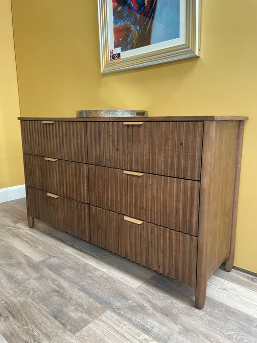 The Cairo Wide Chest, a sustainable wooden dresser with six drawers and vertical grooves, stands against a yellow wall. A round tray sits on top with a framed picture above, all on a light wood floor.