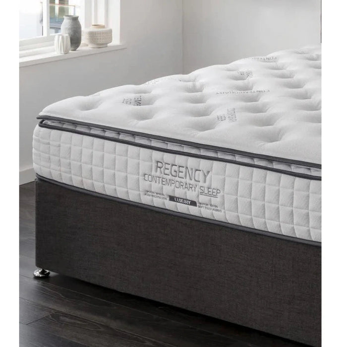 A close-up of a Regency Luxury Mattress by vendor-unknown, featuring a white quilted pillow top on a dark upholstered base in a bright room with a window and decor on the windowsill.