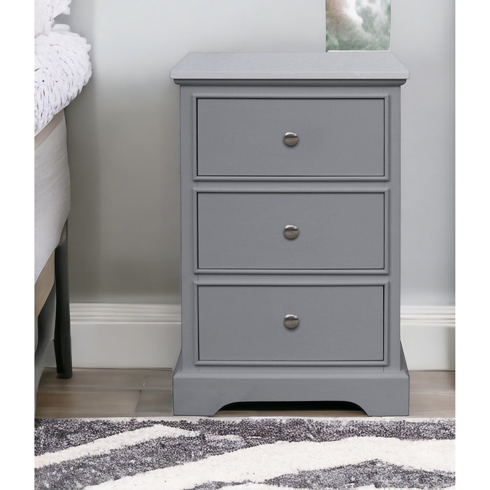 The Russell Light Grey Locker, featuring three drawers with round metal knobs, sits beside a bed and patterned rug on a wood floor, serving as an elegant side table.