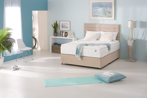 A bright, minimalist bedroom with a beige bed featuring the King Koil - Spinal Pocket Pillow Top mattress, white bedding, and a pastel blue accent wall. A yoga mat and pillow rest on the floor as sunlight streams through sheer curtains.
