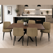 A modern dining area with the Elson Dark Table + 6 Lilly Chairs, black X legs, beige upholstery, and a potted plant centerpiece. A minimalist kitchen with pendant lights, marble backsplash, and light cabinetry is in the background.