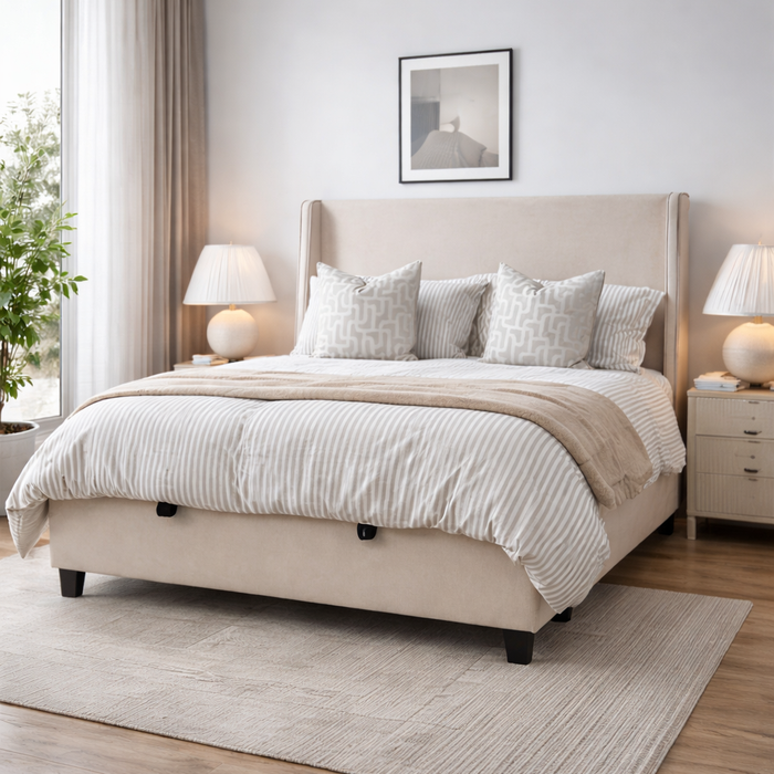 A modern bedroom with a beige linen Rivera Storage Bedframe offering ample storage. Striped bedding and patterned pillows add style, flanked by nightstands with lamps. A framed picture hangs above the bed, and a rug covers the wooden floor.