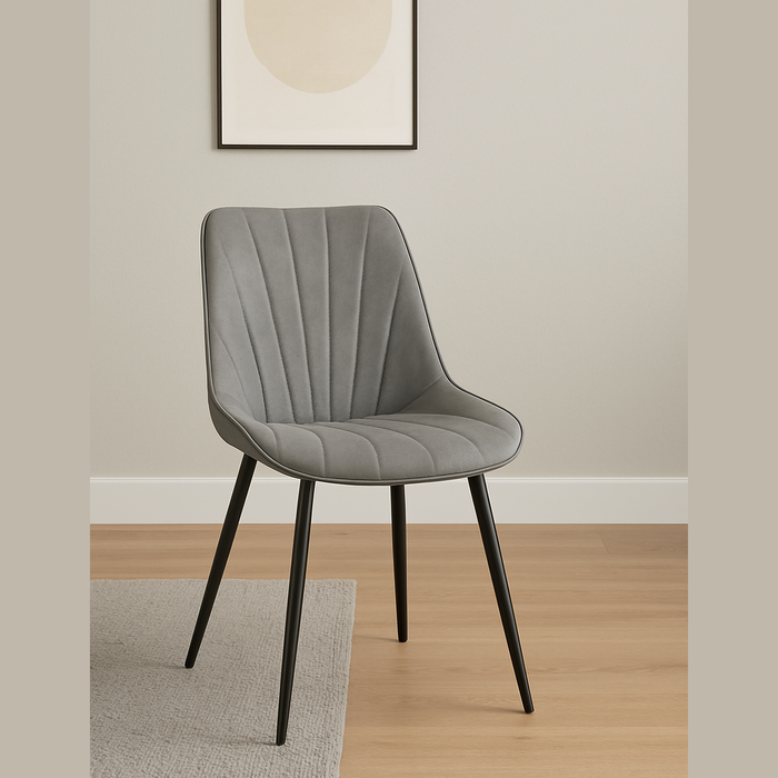 The Lilly Dining Chair, featuring gray upholstery and black metal legs, sits on a light wood floor by a gray rug. Minimalist framed artwork decorates the light-colored wall behind this modern accent piece.