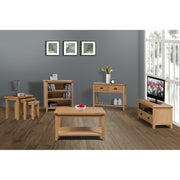 A modern living room with solid oak furniture, including the Saoirse Coffee Table, TV stand, bookshelf, side table set, and console table. Grey walls, wooden floor, three framed pictures, and large windows bring in natural light.