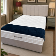 A cozy bedroom features the Fairmont Divan Set - Special with a white mattress and dark blue Fairmont blanket, placed between two lamp-topped bedside tables. Framed botanical prints and an elegant headboard complete the look.