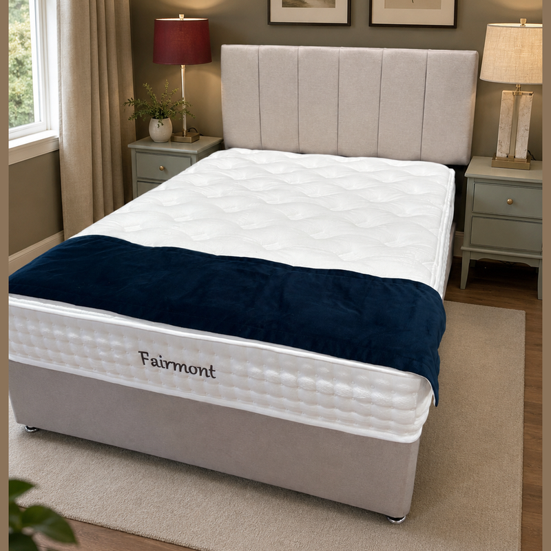A cozy bedroom features the Fairmont Divan Set - Special with a white mattress and dark blue Fairmont blanket, placed between two lamp-topped bedside tables. Framed botanical prints and an elegant headboard complete the look.