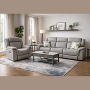 A modern living room featuring a Reno Recliner Sofa in washable fabric, a glass-topped coffee table, light curtains, green plants, wall shelves with decor accents, and a blue and beige area rug on wooden flooring.