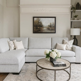 A modern living room featuring the Sorrento Chaise Corner in light gray, neutral pillows, a round coffee table with books and white flowers, wall art above the sofa, decorative shelves, a table lamp, and an indoor plant.