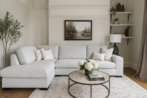 A modern living room featuring the Sorrento Chaise Corner in light gray, neutral pillows, a round coffee table with books and white flowers, wall art above the sofa, decorative shelves, a table lamp, and an indoor plant.
