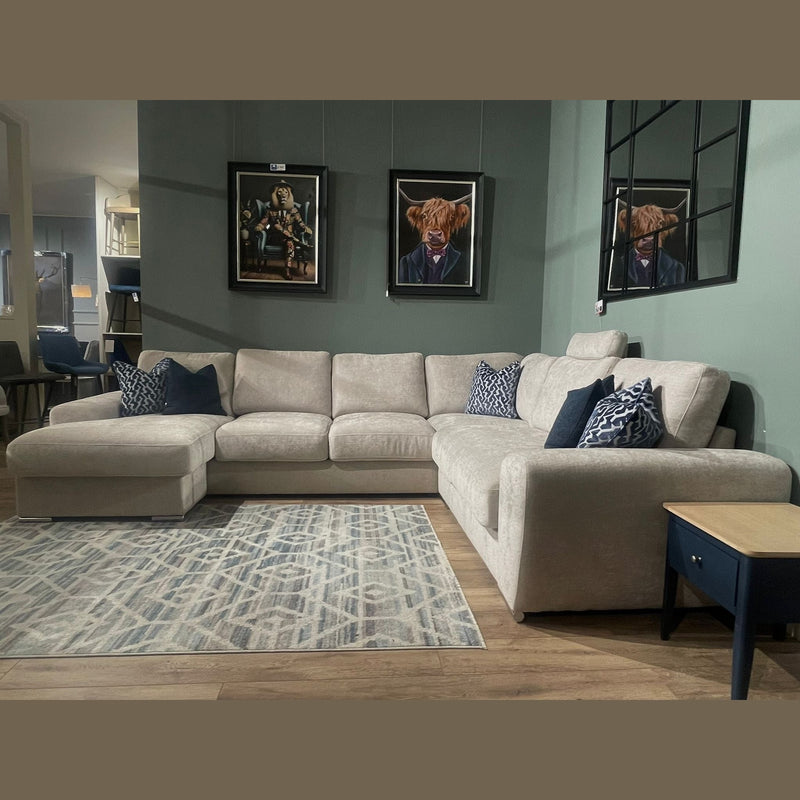 The Grand Corner Group 4L, a large beige corner sofa with blue and patterned pillows, sits on a geometric rug. Behind it are two cow-in-suit paintings and grid-style mirrors on a green wall. A blue side table is nearby.