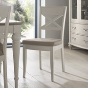 The Montreux Wooden Dining Chair, featuring a white frame, X-shaped backrest, and beige cushioned seat, stands on a light wood floor beside modern dining furniture with large windows overlooking a lush green garden.