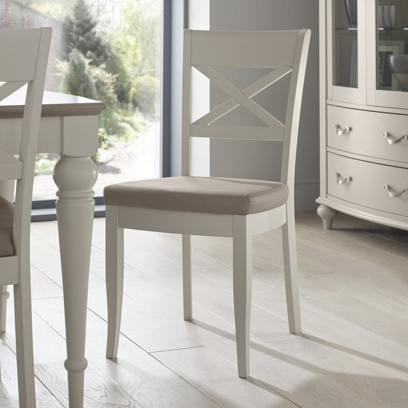 The Montreux Wooden Dining Chair, featuring a white frame, X-shaped backrest, and beige cushioned seat, stands on a light wood floor beside modern dining furniture with large windows overlooking a lush green garden.