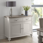 The Montreux Small Sideboard in grey washed oak, with two drawers and two cabinets, stands against a wall. Its top displays a silver lamp, white tulips in a vase, and a fruit plate. Large windows show greenery outside.