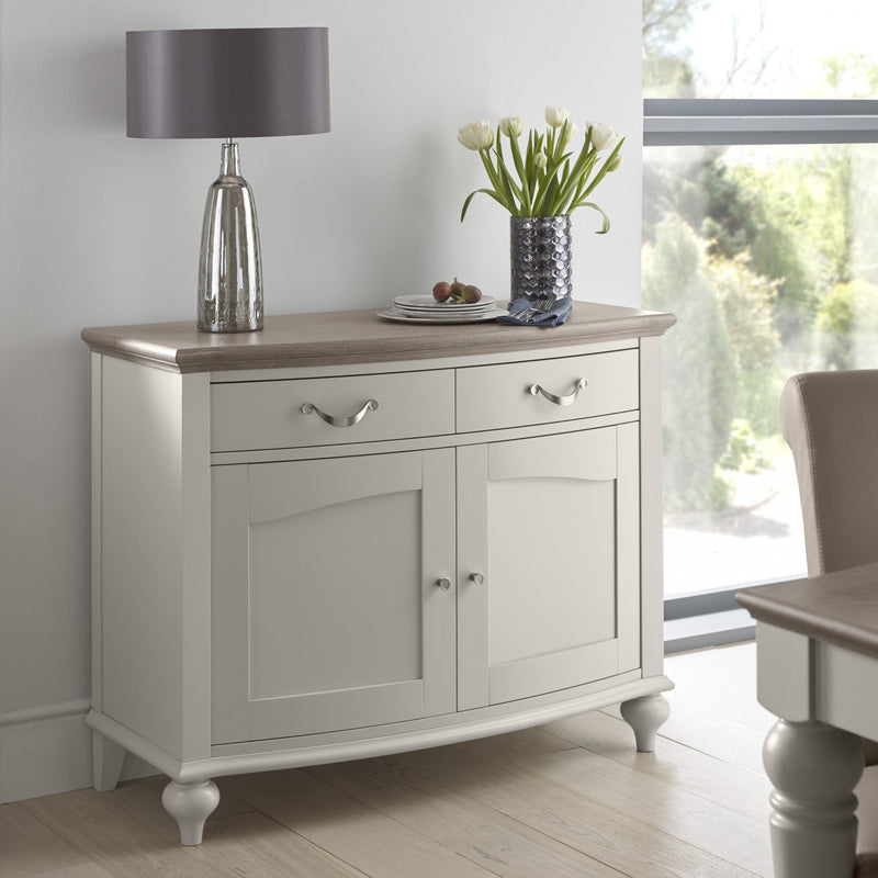 The Montreux Small Sideboard in grey washed oak, with two drawers and two cabinets, stands against a wall. Its top displays a silver lamp, white tulips in a vase, and a fruit plate. Large windows show greenery outside.