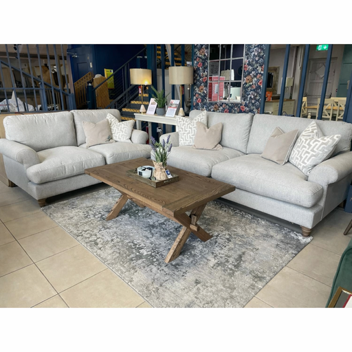 A modern living room features two light gray sofas from the Monroe Sofa Range, accented with beige and patterned Irish fabric cushions. A wooden coffee table, gray rug, decorative items, floor lamps, and a floral wall complete the look.