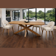 A modern dining room showcases the Ashton Dining Table in solid ash wood, paired with four cushioned chairs on a dark wood floor. Large windows offer views of a green outdoor space with trees and patio furniture.
