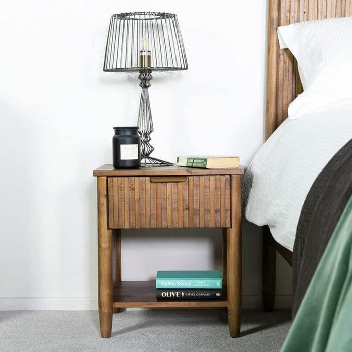 Wooden nightstand with lamp and books next to a bed.