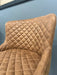 A close-up of the Diamond Dining Chair, featuring brown upholstery with a quilted pattern on the backrest and seat, set against a light blue wall.
