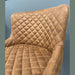 A close-up of the Diamond Dining Chair, featuring brown upholstery with a quilted pattern on the backrest and seat, set against a light blue wall.