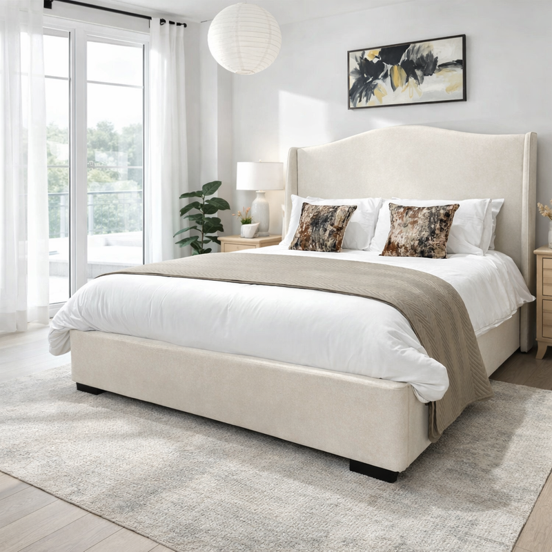 Modern bedroom with the Venice Bedframe 5ft in a cream upholstered finish featuring a curved headboard, white bedding, decorative pillows, wooden nightstands, a table lamp, a plant, large windows with sheer curtains, and abstract artwork above the bed.