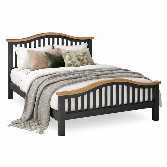 The Capri Dark Bedframe in black wood with curved smokey oak accents is styled with white bedding, assorted neutral and green pillows, and a gray knit throw draped at the foot of the bed.
