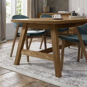 A modern dining room with the Camden Dining Table, teal cushioned chairs, a light patterned rug on wooden floors, and large windows filling the space with natural light. A cup, saucer, and black pitcher sit on the extending table.