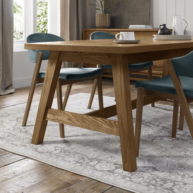 A modern dining room with the Camden Dining Table, teal cushioned chairs, a light patterned rug on wooden floors, and large windows filling the space with natural light. A cup, saucer, and black pitcher sit on the extending table.