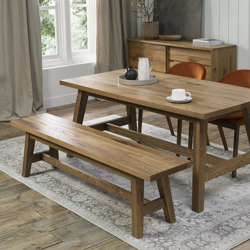 A modern dining room features an oak table, Camden Bench, and two orange mid-century armchairs. A white rug lies on the wood floor, with large curtained windows and a sideboard completing the inviting setting.